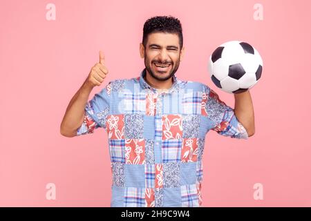 Happy excited bearded man with dark hair and beard wearing blue shirt posing with soccer ball and showing thumb up, cheering on football match. Indoor studio shot isolated on pink background. Stock Photo