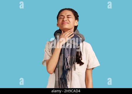 Unhappy sick woman wrapped in scarf, touches neck, suffers from sore throat, cant breath well, chokes and smirks face, feels discomfort and suffocation. Indoor studio shot isolated on blue background. Stock Photo