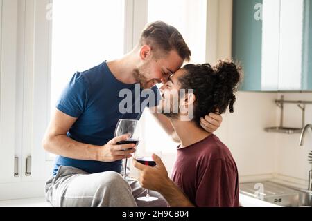 Tenderness moment of smiling gay couple, young man toasting with red wine in the kitchen, male couple having fun and drinking red wine for celebration Stock Photo