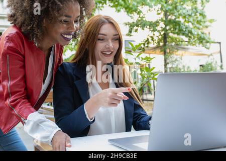 Two business women analyzing documents on laptop while sitting on a table in outdoors workplace. Woman executives at work showing some achievement to Stock Photo