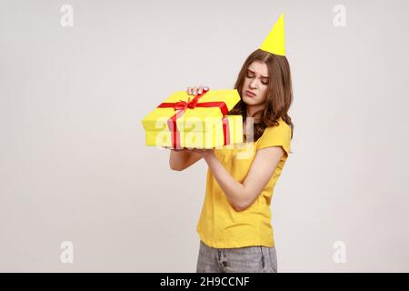 Upset teenager girl opening birthday gift box and looking inside with disappointed expression, unwrapping bad present, wearing yellow T-shirt and birthday cone. Indoor isolated on gray background. Stock Photo
