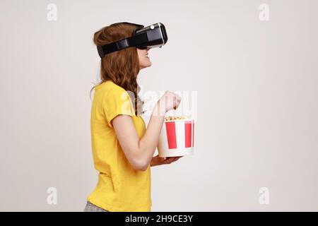 Profile portrait of young brown haired woman in VR headset watching movie with popcorn, wearing yellow casual t-shirt, eating tasty snack. Indoor studio shot isolated on gray background. Stock Photo