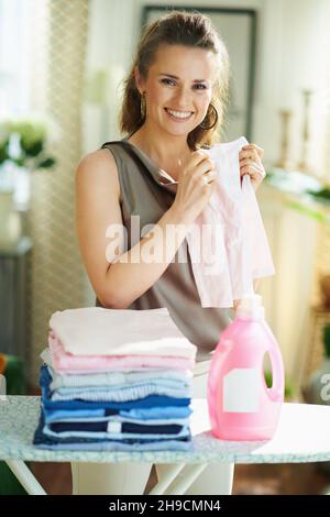 happy modern woman in silk blouse and beige pants with ironing board, pile of folded ironed clothes and pink bottle of laundry detergent at modern hom Stock Photo