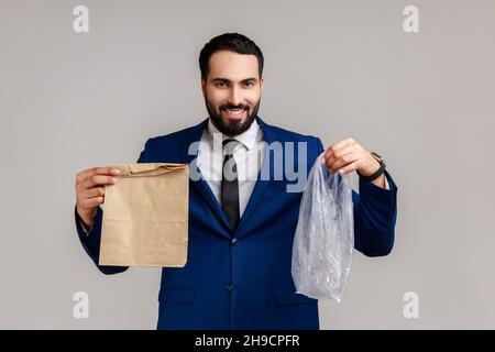 Smiling businessman holding out hands with paper and poly bag, giving you chance to make right choice and save planet, wearing official style suit. Indoor studio shot isolated on gray background. Stock Photo