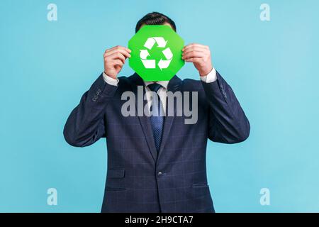 Unknown male person wearing official style suit hiding face behind green recycling sign, garbage sorting and environment protection, thinking green. Indoor studio shot isolated on blue background. Stock Photo