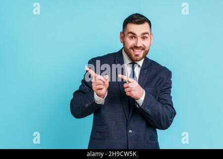 Advertise here. Positive bearded man wearing official style suit pointing finger away paying your attention at empty space for advertisement. Indoor studio shot isolated on blue background. Stock Photo