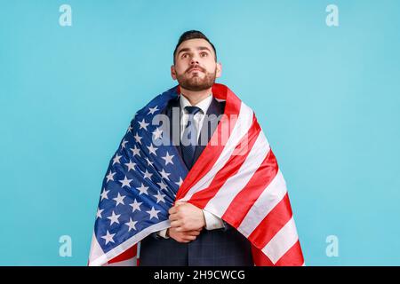 Young adult bearded businessman being wrapped in USA flag, celebrating labor day or US Independence day 4th of july, government employment support. Indoor studio shot isolated on blue background. Stock Photo