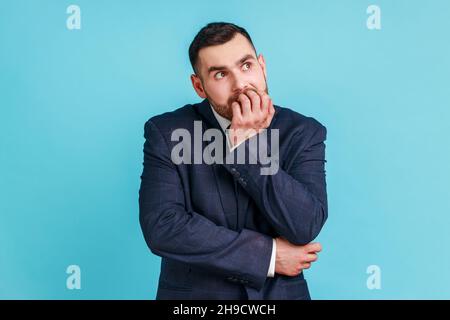 Anxious nervous businessman with beard in official style suit biting nails on fingers looking at camera with terrified expression, worried about work. Indoor studio shot isolated on blue background. Stock Photo
