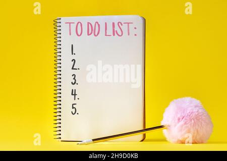 Open spiral notebook with to do list and golden pen with pink fluffy ball on bright yellow background Stock Photo