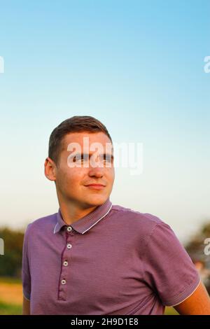 Young man. Portrait of smiling young brunette man in purple polo shirt standing outdoor on nature background at summer day. 20s years guy. Millennial Stock Photo