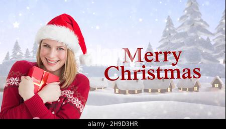 Composite image of happy young woman in santa hat embracing gift by christmas greeting Stock Photo
