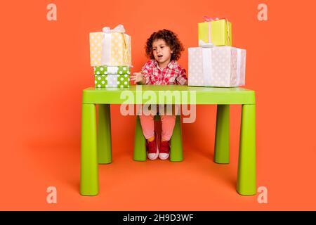 Photo of moody upset schoolgirl wear plaid outfit sitting green table dislike gifts boxes isolated orange color background Stock Photo