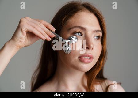 Young white woman using face massager while standing isolated on gray background Stock Photo
