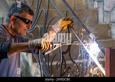Man wearing protective glasses welds metal with welding machine. Sparks from welding fly to sides. Close-up Stock Photo