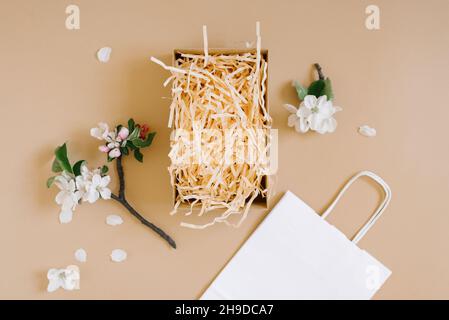 Mockup of an open gift box with peach colored paper shavings, white paper bag and apple flowers on a beige background. Flat lat, top viw Stock Photo