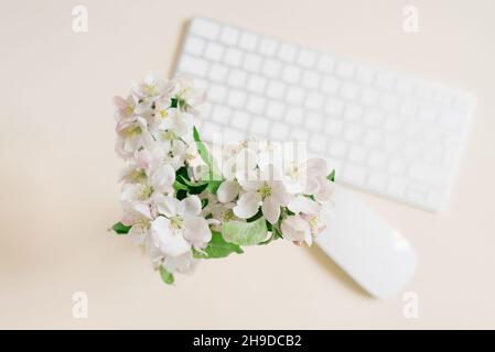 A white vase with a beautiful bouquet of branches of a blossoming apple tree on a beige table and a keyboard with a mouse in decoloration, top view. Stock Photo