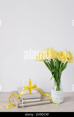 A gift box with a bow in the workplace with bouquets of bright yellow flowers and a stack of notebooks or books. Flowers for a business woman. A gift Stock Photo