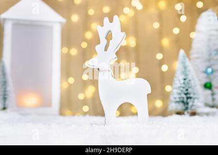Christmas fairy tale composition with white wooden deer toy, lantern and small Christmas trees on the snow against warm lights bokeh background. Merry Stock Photo