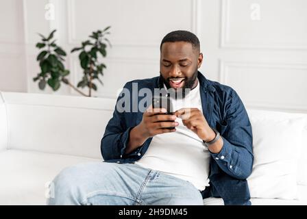 Excited young African-American bearded guy in casual using smartphone sitting on the sofa at modern apartment, cheerful black guy scrolling news feed, messaging in social networks, using dating app Stock Photo