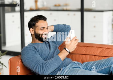 Hilarious 30s Indian man using smartphone sitting on the couch at home, Hispanic latin male holds mobile phone and laughing cheerfully, watching comedy, funny videos, joking with friends online Stock Photo