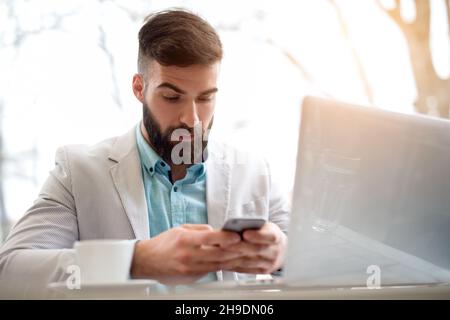 Handsome bearded man in stylish white and blue suit works on computer checking emails on smart phone. Stock Photo