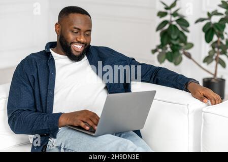 Serene young 30s African-American guy using laptop in relaxed home atmosphere, lying on the sofa and typing, web surfing, watching movies, carefree black man spends leisure online Stock Photo