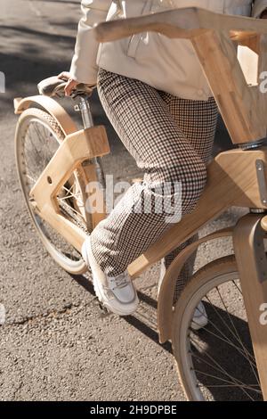 From above unrecognizable woman in checkered pants taking seat on wooden eco bicycle during ride on city street. Stock Photo