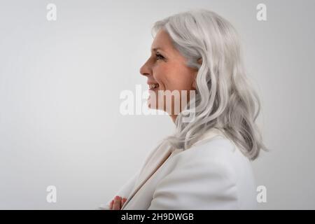 Studio shot of grey haired senior woman wearing elegant clothes while standing at isolated grey background. Copy space. Side view. Stock Photo
