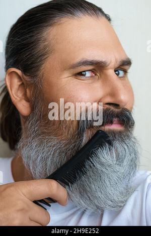 Close-up portrait of caucasian middle-aged man with hairbrush. He comb his grey beard. Selective focus.  Stock Photo