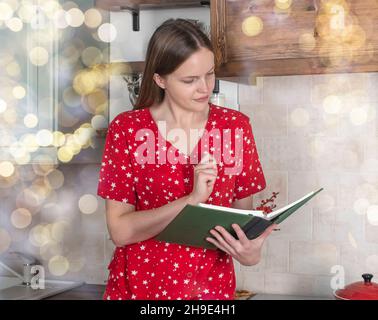 Young woman reading recipe book at kitchen interior. Modern female in pajamas with cookbook at home. Stock Photo