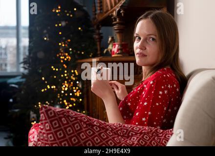 Young woman portrait with hot tea cup sitting in chair at hygge Scandi home on winter holidays with fir tree and lights. Stock Photo