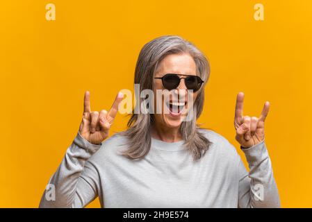 Crazy positive sanguine senior woman in sunglasses is showing sign of horns, funny hilarious mature modern gray-haired lady making symbols of rock standing isolated over yellow background Stock Photo