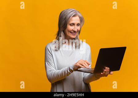 Confident modern mature woman using laptop isolated on yellow, senior female entrepreneur typing, middle-aged lady with gray hair in casual wear checking emails Stock Photo