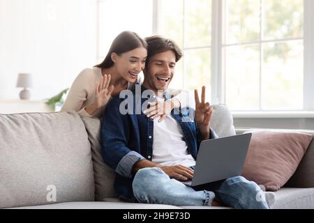 Smiling millennial european couple communicate with family remotely, wave hand, showing peace sign Stock Photo