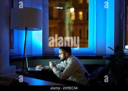 Tired web designer spend night at work before project launch working on laptop and drinking coffee Stock Photo