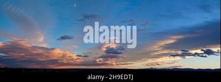 The setting sun highlights clouds just above the horizon and below the early evening half moon. Stock Photo