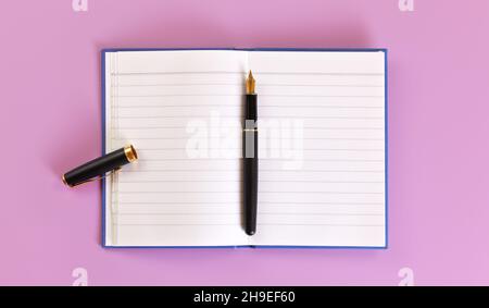 Top down view - black fountain ink pen with gold nib, opened cap, empty white paper pad on lilac board under Stock Photo