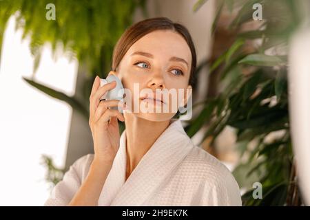 Portrait of beautiful young woman with flawless skin using silicone face brush, standing in bathroom decorated with green plants Stock Photo