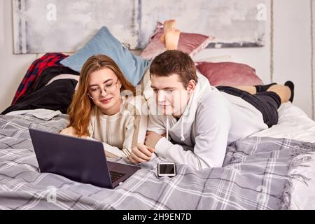 Online shopping using laptop for young couple lying on their stomachs in bed in their bedroom. Stock Photo