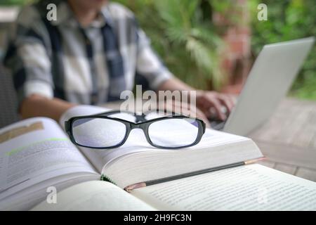 Books on table with woman and computer laptop at the background. Selective focus on eyeglasses. Stock Photo