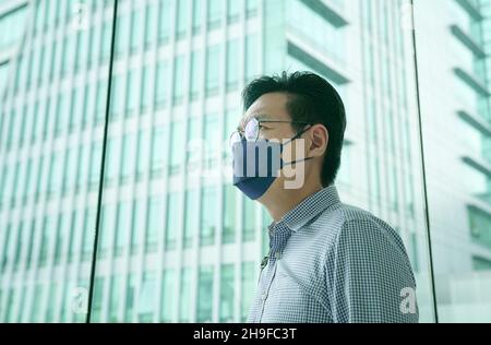 Asian businessman wearing face mask with office building at the background. Business during Covid-19 pandemic concept. Copy space. Stock Photo