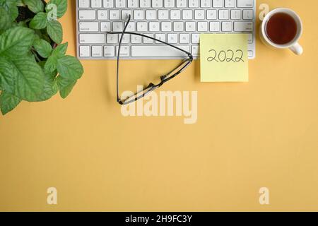 2022 written on yellow paper stick on top of keyboard with coffee, eyeglasses and plant. Flat lay, copy space. Stock Photo