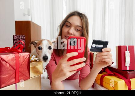 Woman buys gifts online through a smartphone with her pet dog sitting at a table among gifts packed boxes in colored paper with bows and ribbons at ho Stock Photo