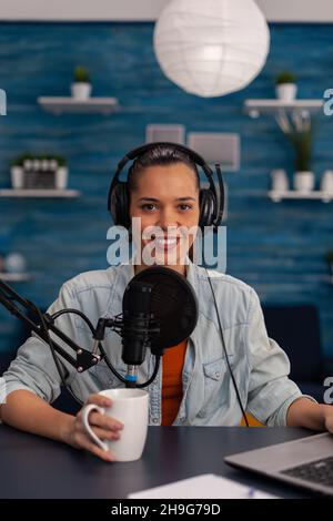 Portait of woman vlogger holding cup and streaming podcast using laptop. Social media influencer recording livestream with headphones and microphone in home office. Smiling blogger podcasting. Stock Photo