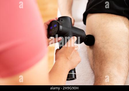 Massage gun background. Young female physiotherapist using handheld massaging gun to relieve leg muscle pain during physical therapy session.