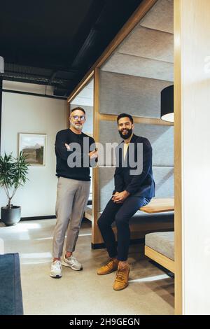 Two successful businessmen looking at the camera while standing in an office. Two cheerful businessmen working together in a modern co-working space. Stock Photo