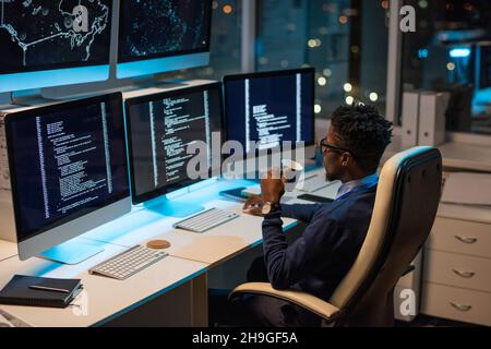 Young African programmer with drink looking at coded data on computer screen while working in office Stock Photo