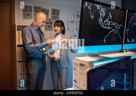 Two intercultural colleagues discussing paper while standing by whiteboard with business documents in office Stock Photo