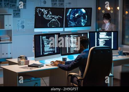 Young contemporary cyber security manager typing while sitting by desk in front of computer monitors Stock Photo