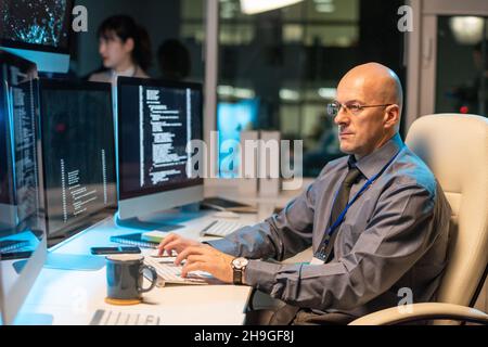 Serious bald businessman looking through data on computer screen while sitting in front of monitor by desk Stock Photo